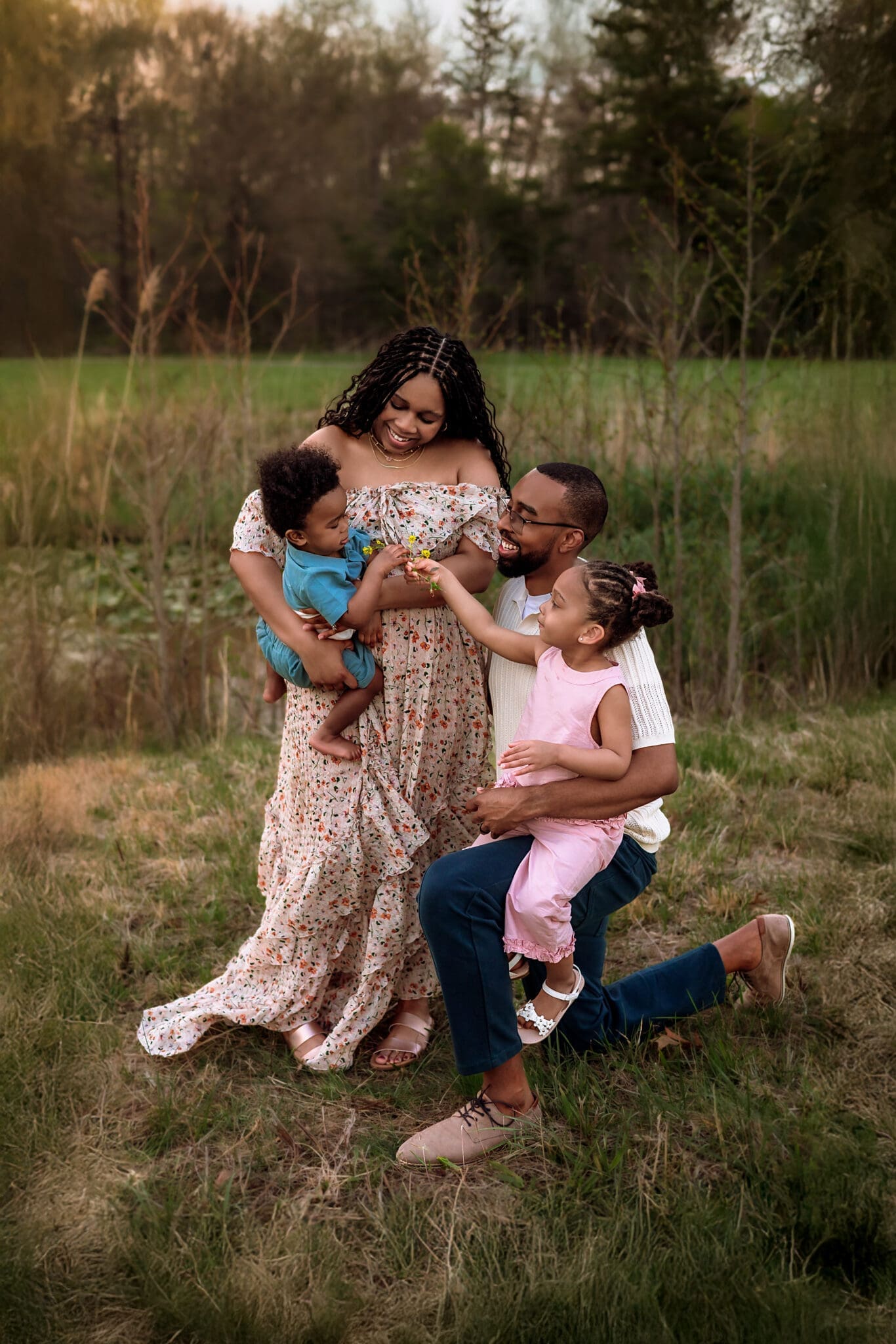 A photo of a smiling family of four sharing a playful moment together in a grassy field, with parents holding their young children on a warm evening. Taken by Erin Link, Maryland Family Photographer in Bowie.