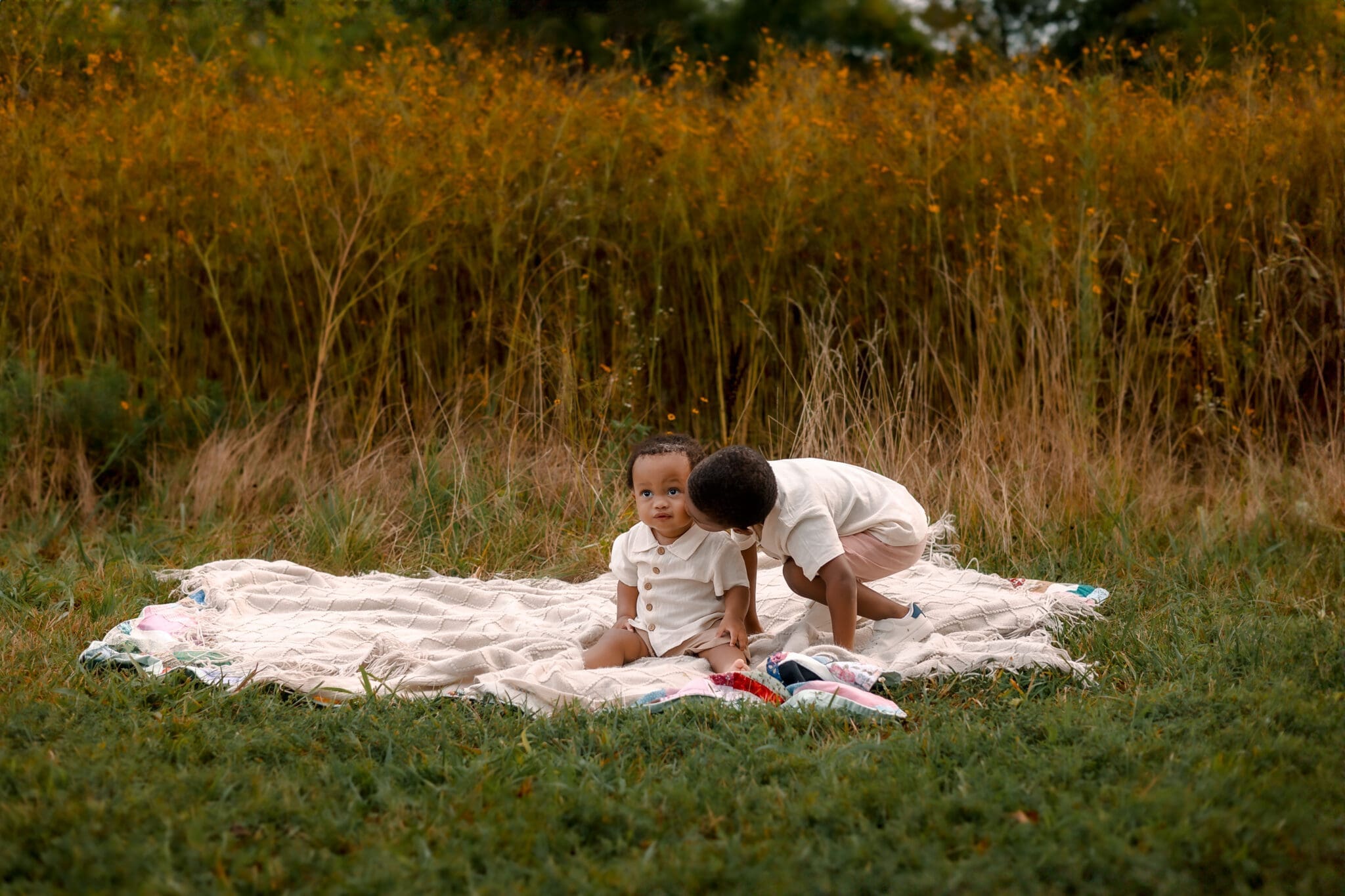 Brother kissing cheek of Child sitting on blanket in a tall grass meadow during family session