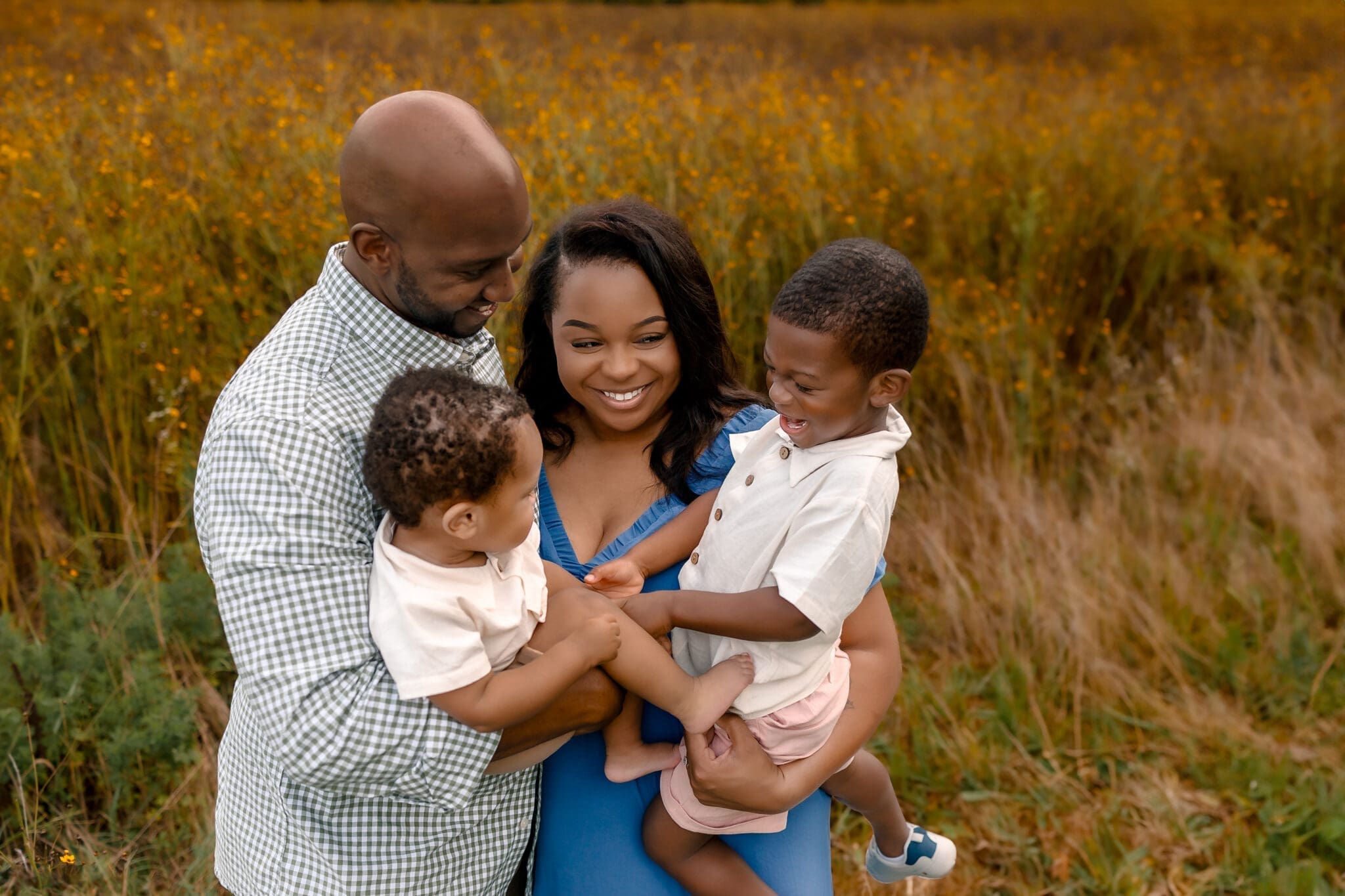 Parents holding two toddlers and smiling together in tall grass at golden hour