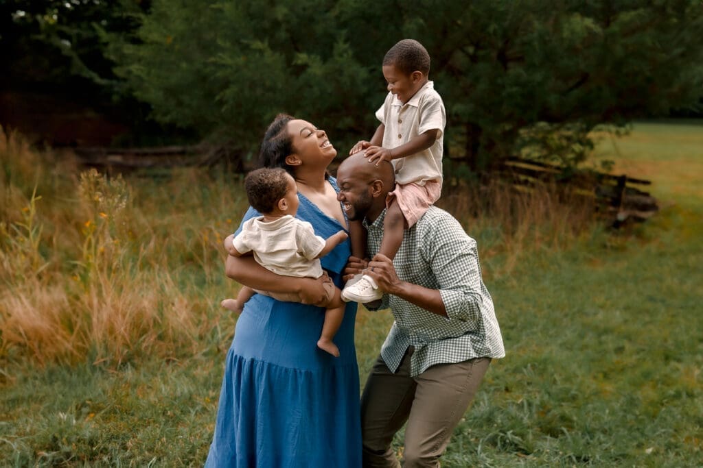 A family of four stands together in tall grass during a summer lifestyle session, laughing and snuggling under warm evening light in Maryland.