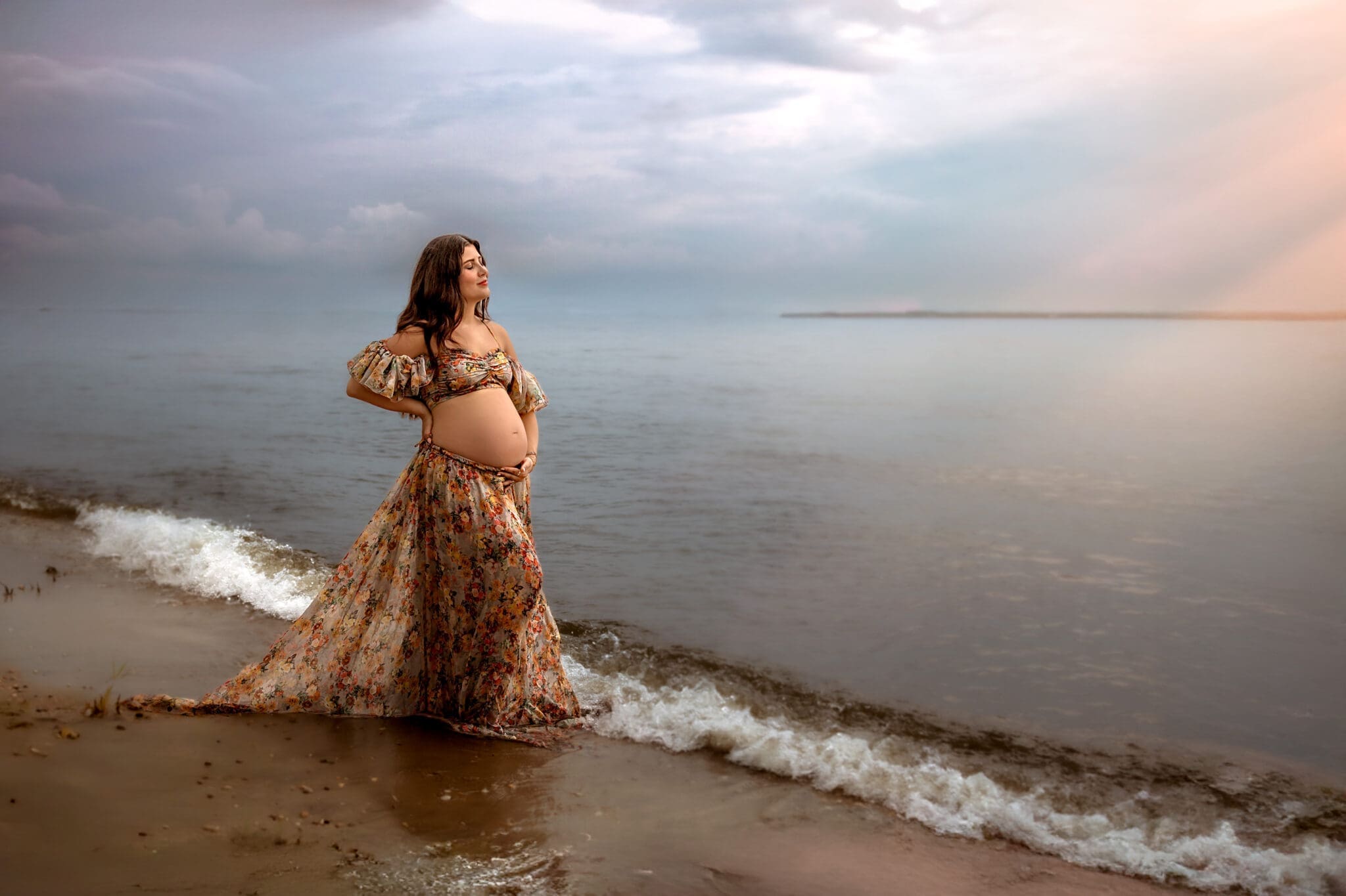 Pregnant woman in a flowing dress stands at the water’s edge during a maternity photography session in Annapolis, MD.