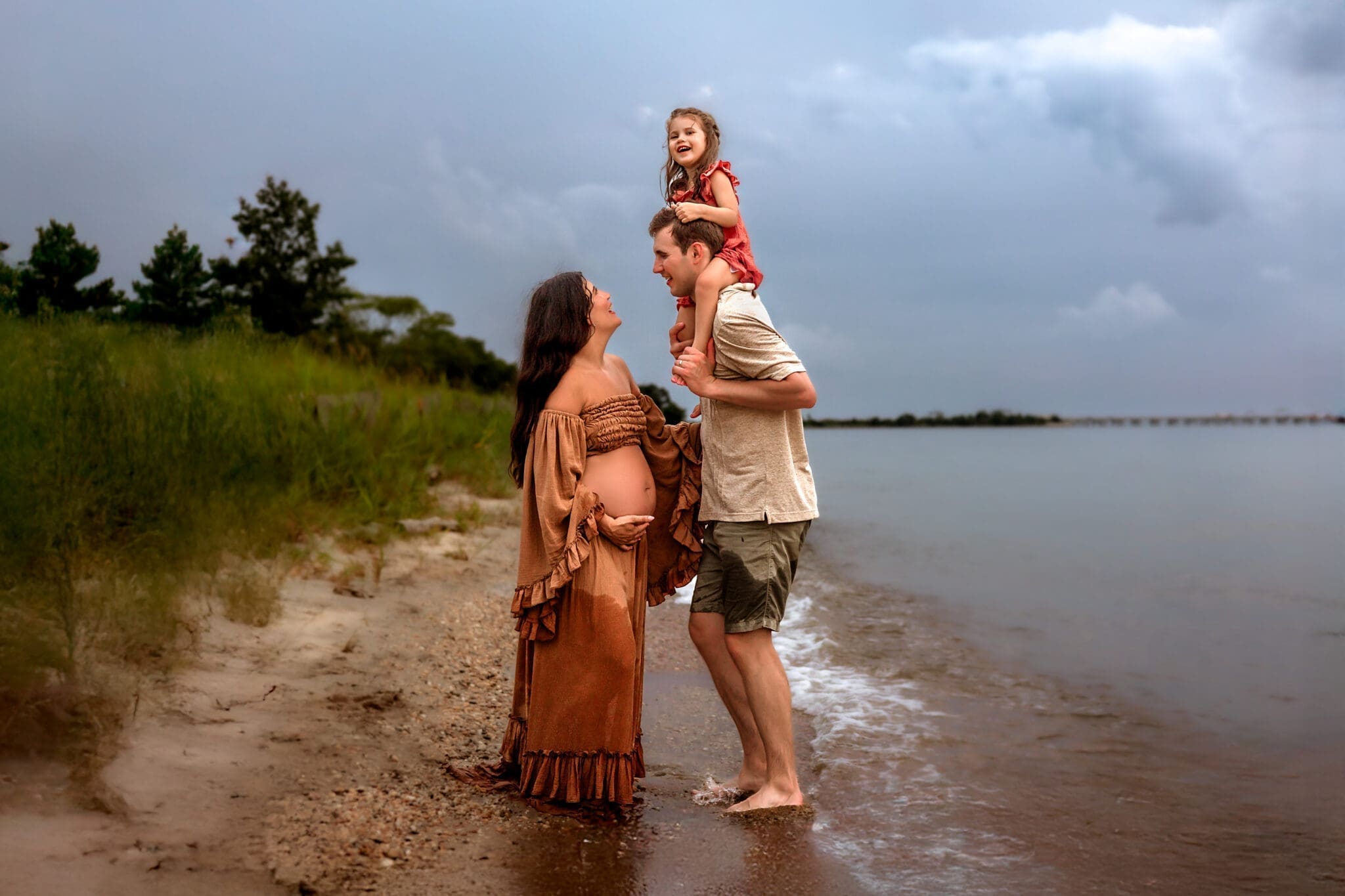 A pregnant woman in a rust-colored dress stands on the shoreline, smiling at her partner who holds their young daughter on his shoulders. The family is barefoot at the water’s edge with cloudy skies in the background, creating a tender and joyful beach moment.