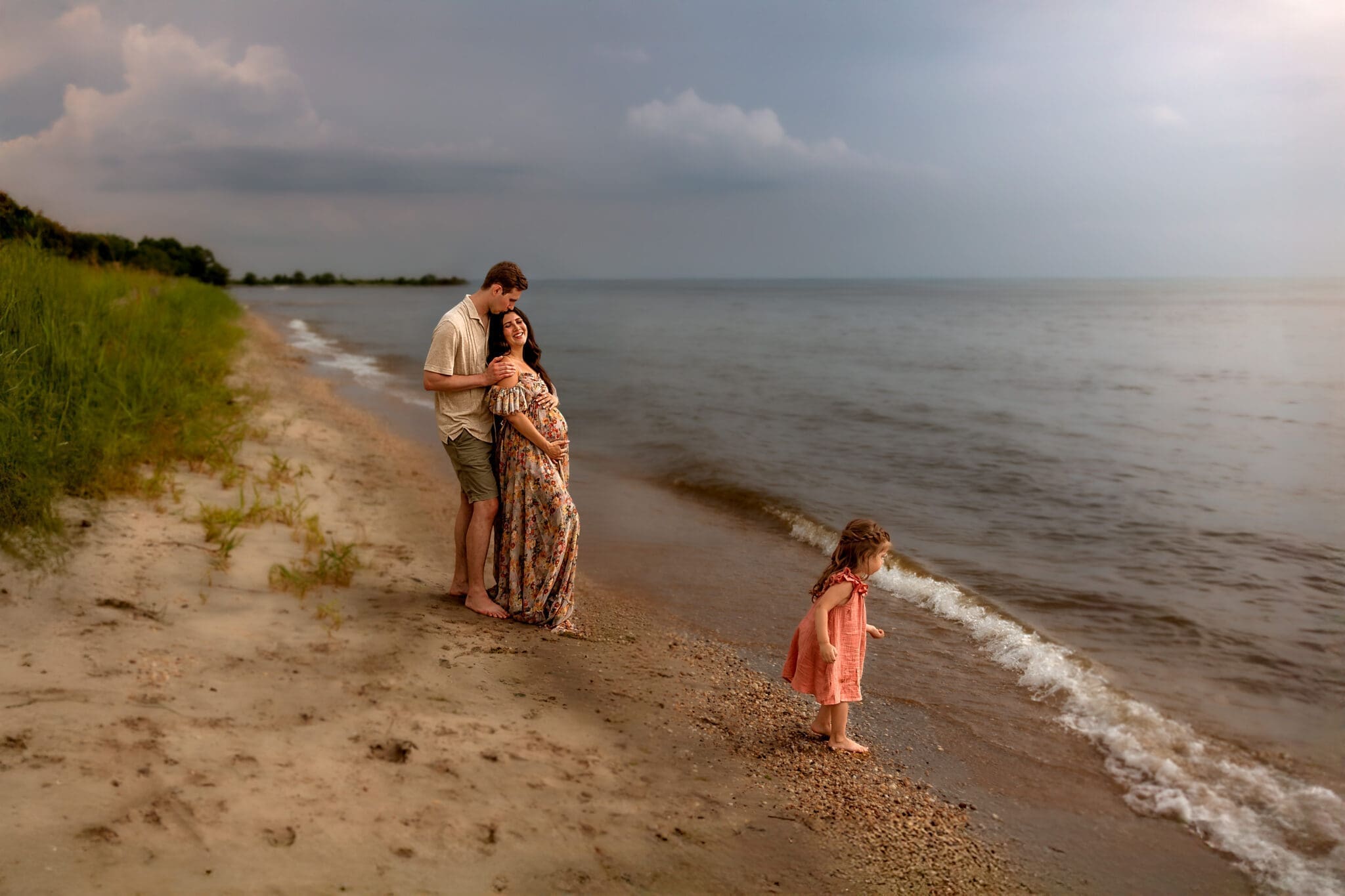 Family walks along the shoreline during an Annapolis beach maternity photo session, with golden light reflecting on the water.