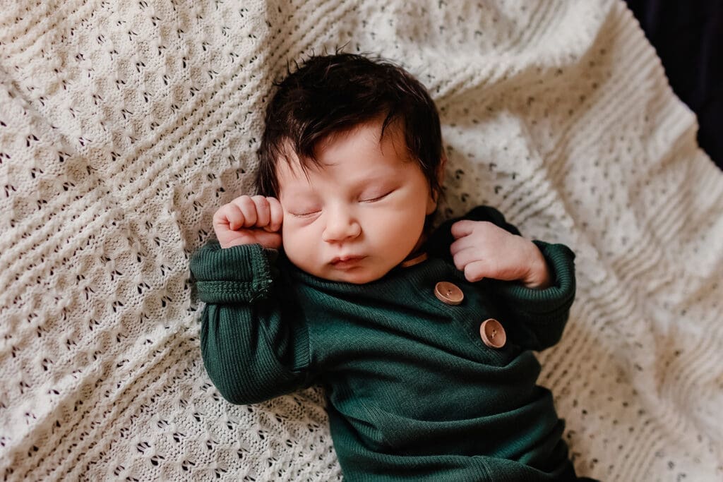 Newborn baby in a forest green romper sleeping peacefully on a textured blanket during an in-home session.