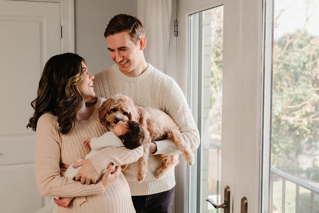 Parents hold their baby near a large window with their dog in the foreground during an in-home newborn session in Bethesda, Maryland.