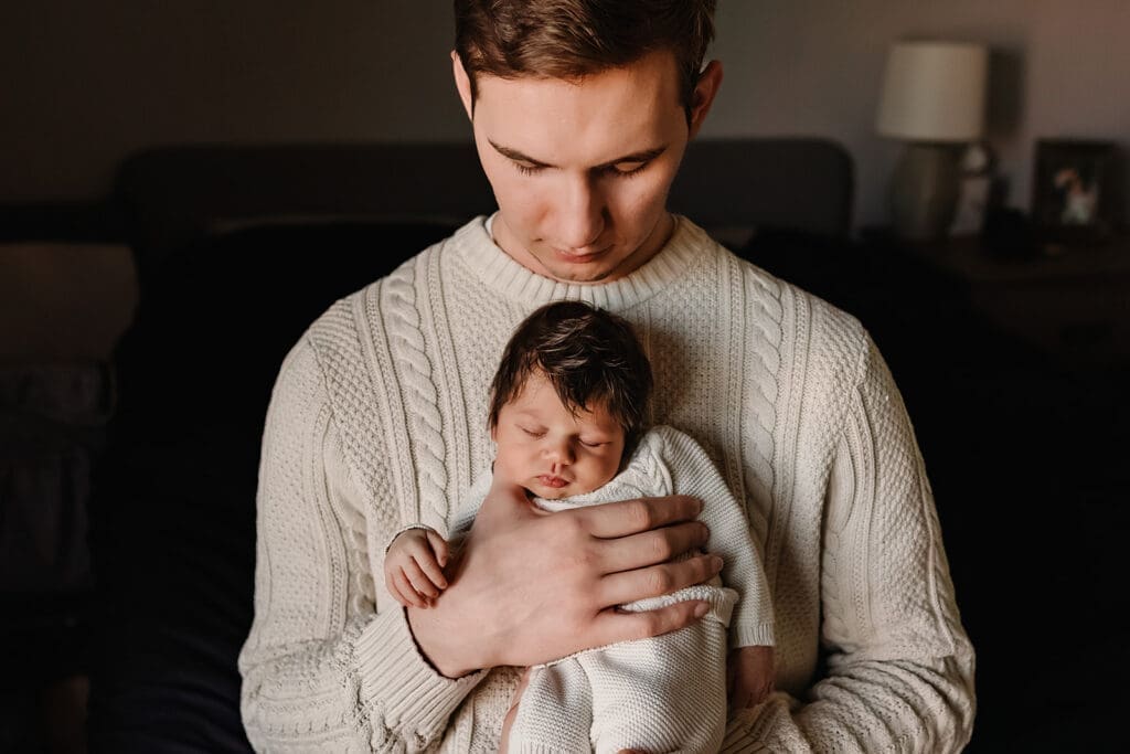 Dad holds his swaddled newborn close to his chest during a quiet moment in an in-home newborn photography session.