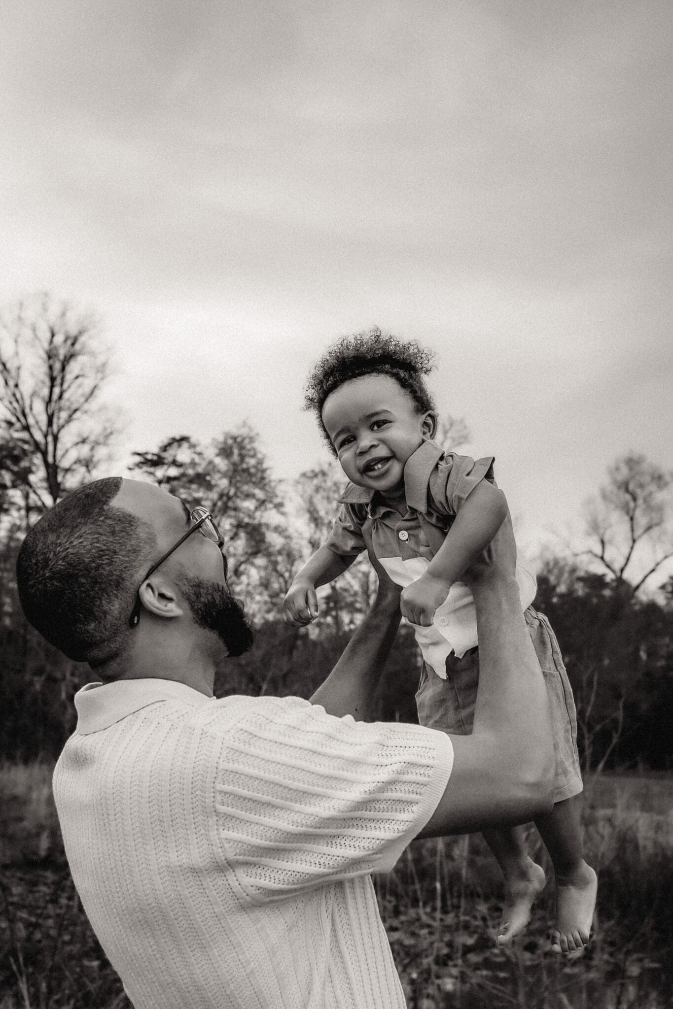 A parent lifts their toddler high in the air during a black-and-white lifestyle family photography session in Maryland, with a pond in the background.