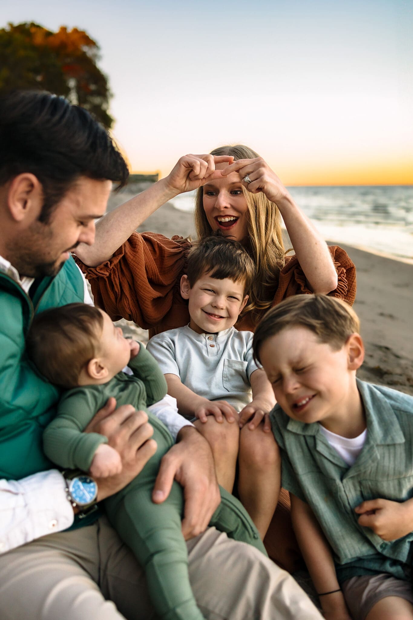 The family sits together on a soft blanket at sunset, dad holding the baby, mom sings Itsy Bitsy Spider with her hands, and two big brothers curled up beside them.