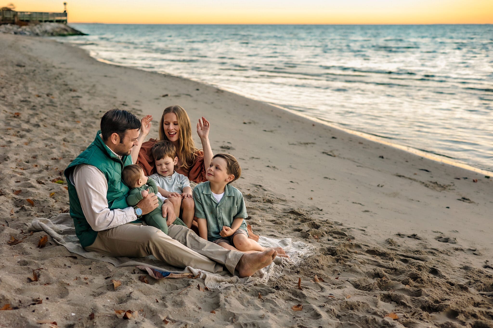 The family sits together on a soft blanket at sunset, dad holding the baby, mom playfully framing the scene with her hands, and two big brothers curled up beside them.