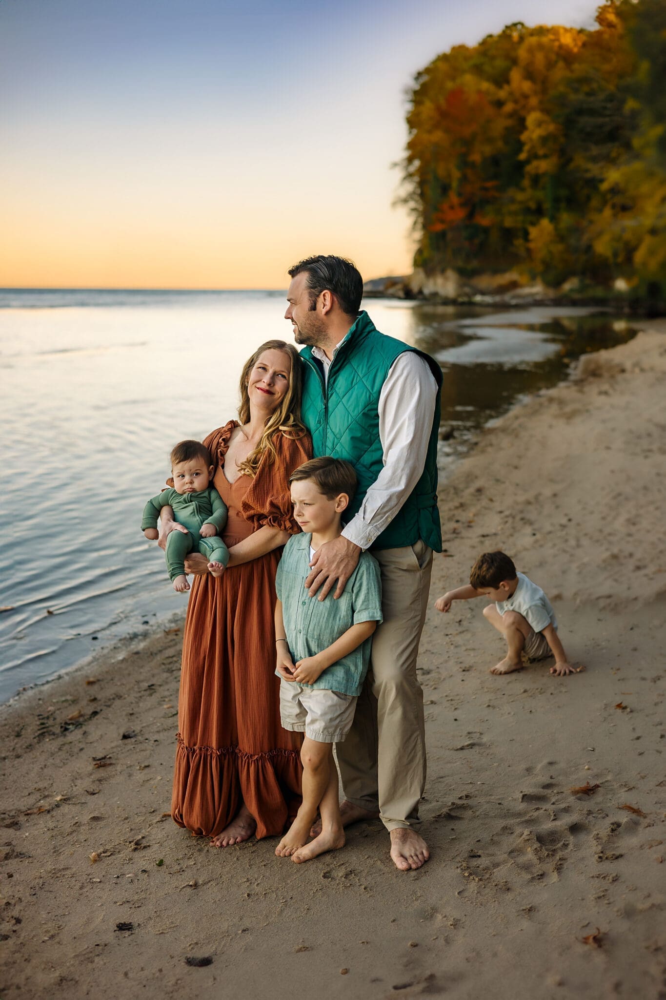 Parents stand at the water’s edge, arms around each other and one boy in front, as another sibling digs in the sand nearby.