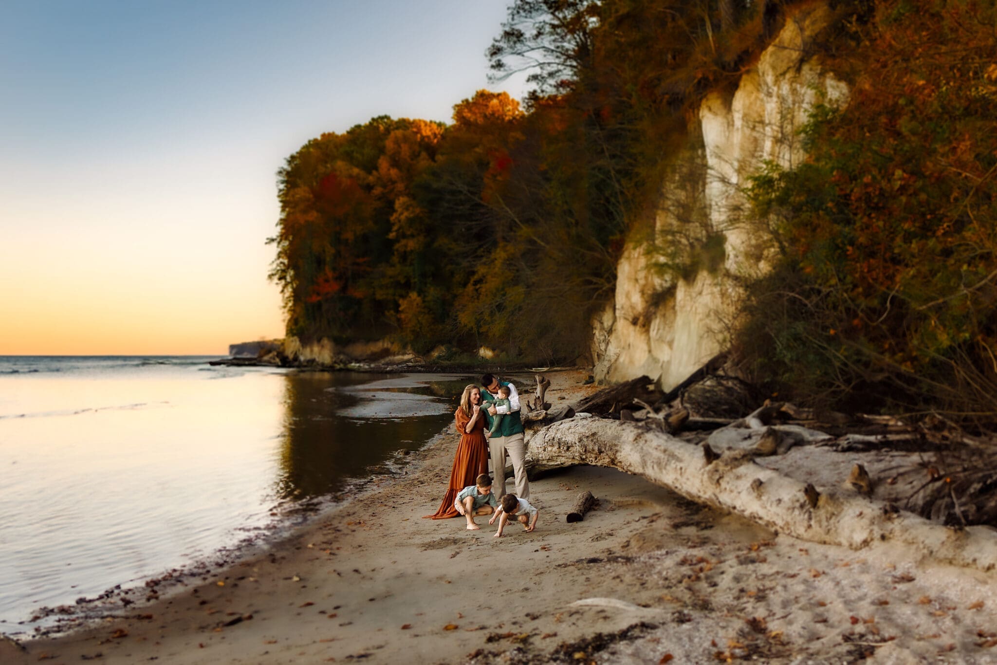 A wide shot shows a family clustered by a driftwood log under white cliffs draped in fall foliage, with the boys exploring the sand at their feet.