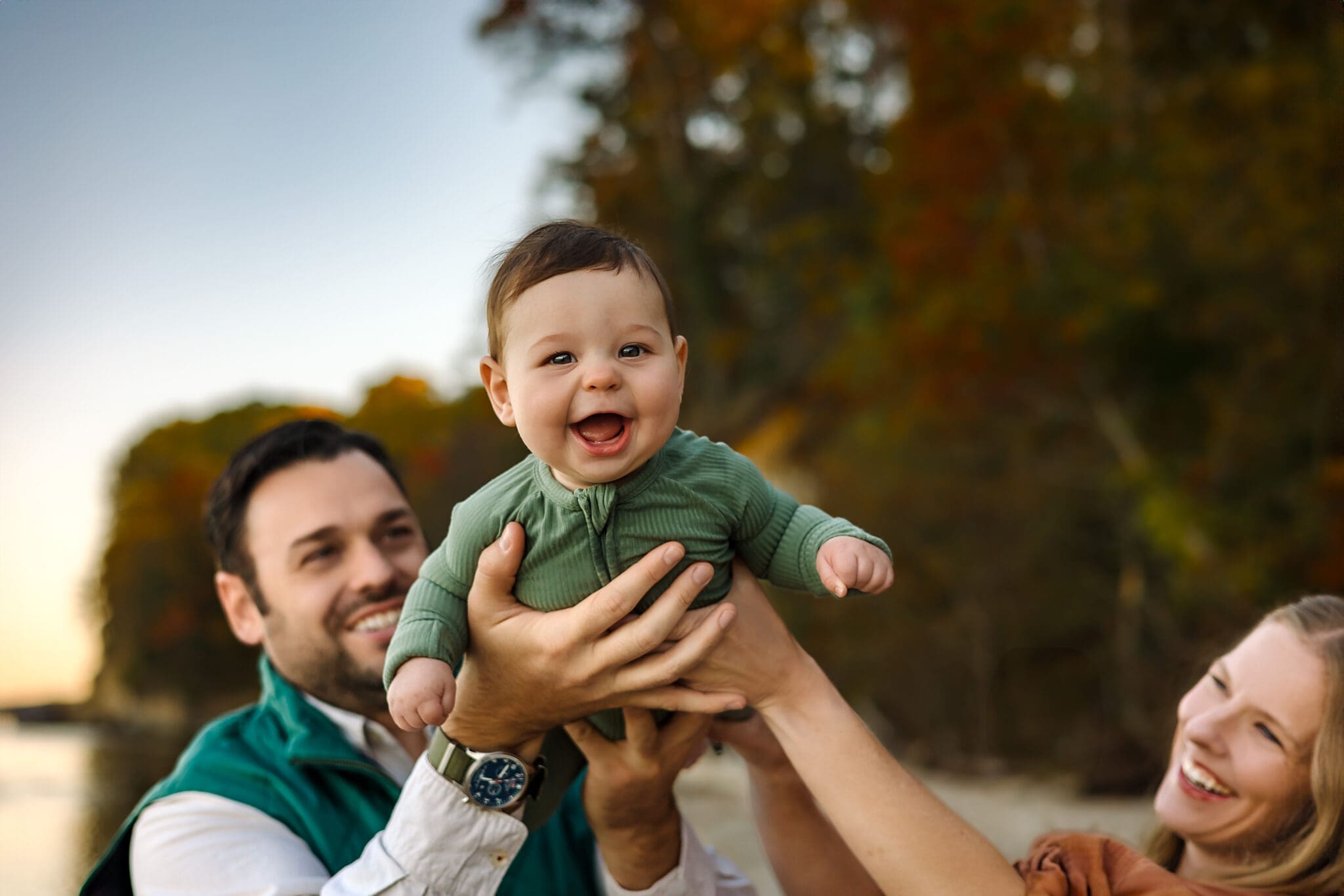 Dad and mom’s hands cradle the baby high above their heads, the baby’s face lighting up against a backdrop of colorful trees and calm bay water.