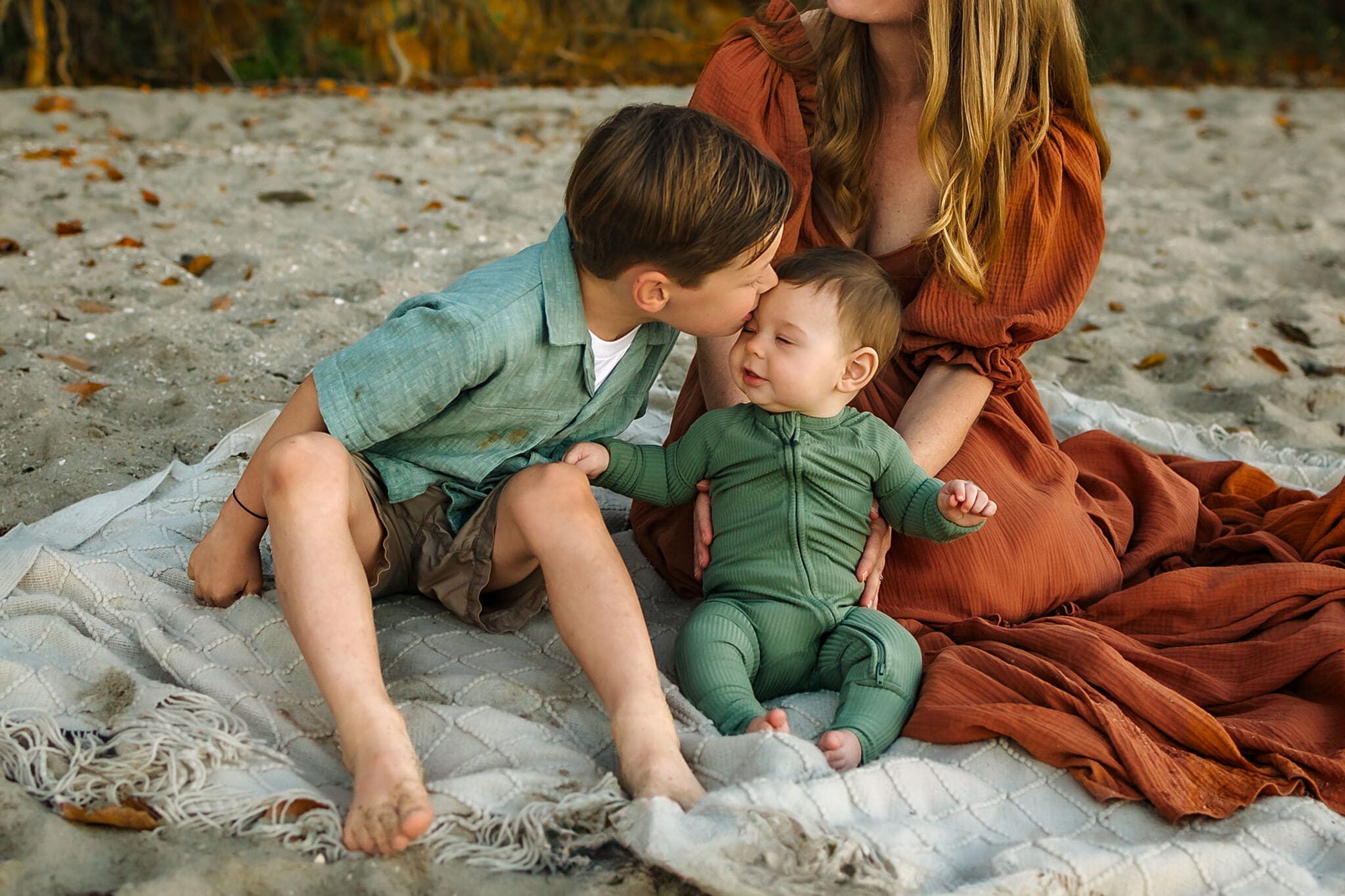 An older brother leans in to kiss his little sibling’s forehead as the beach waves lap softly behind them.