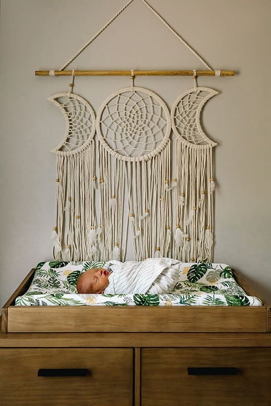 Newborn swaddled in a light blanket on a wooden changing table in a Bethesda, Maryland home, with a leafy-patterned cover and macramé triple moon wall hanging in the background during an in-home newborn photography session.