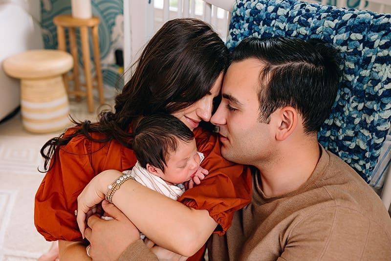Mother in rust-colored blouse and father in brown sweater cradle their newborn, sharing a quiet moment at home in Bethesda, Maryland.