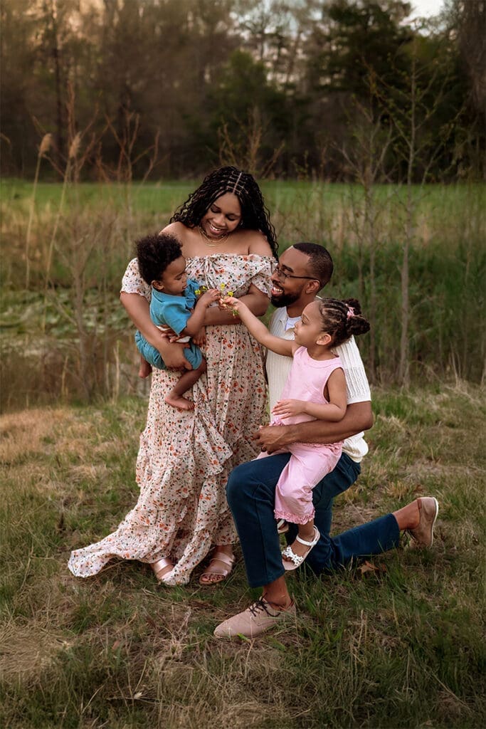 A photo of a smiling family of four sharing a playful moment together in a grassy field, with parents holding their young children on a warm evening. Taken by Erin Link, Maryland Family Photographer in Bowie.