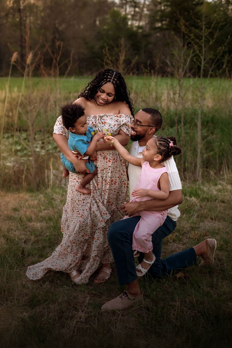 A photo of a smiling family of four sharing a playful moment together in a grassy field, with parents holding their young children on a warm evening. Taken by Erin Link, Maryland Family Photographer in Bowie.