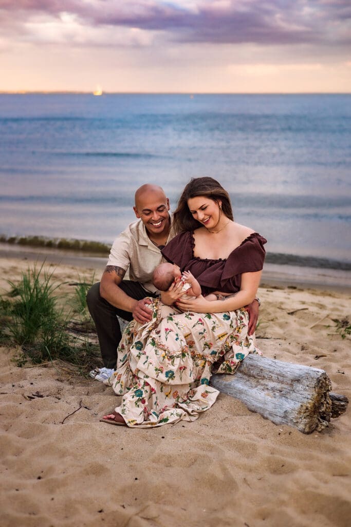 Happy family of three sitting on driftwood at Maryland beach during golden hour sunset - parents smiling while holding newborn baby, ocean waves in background
