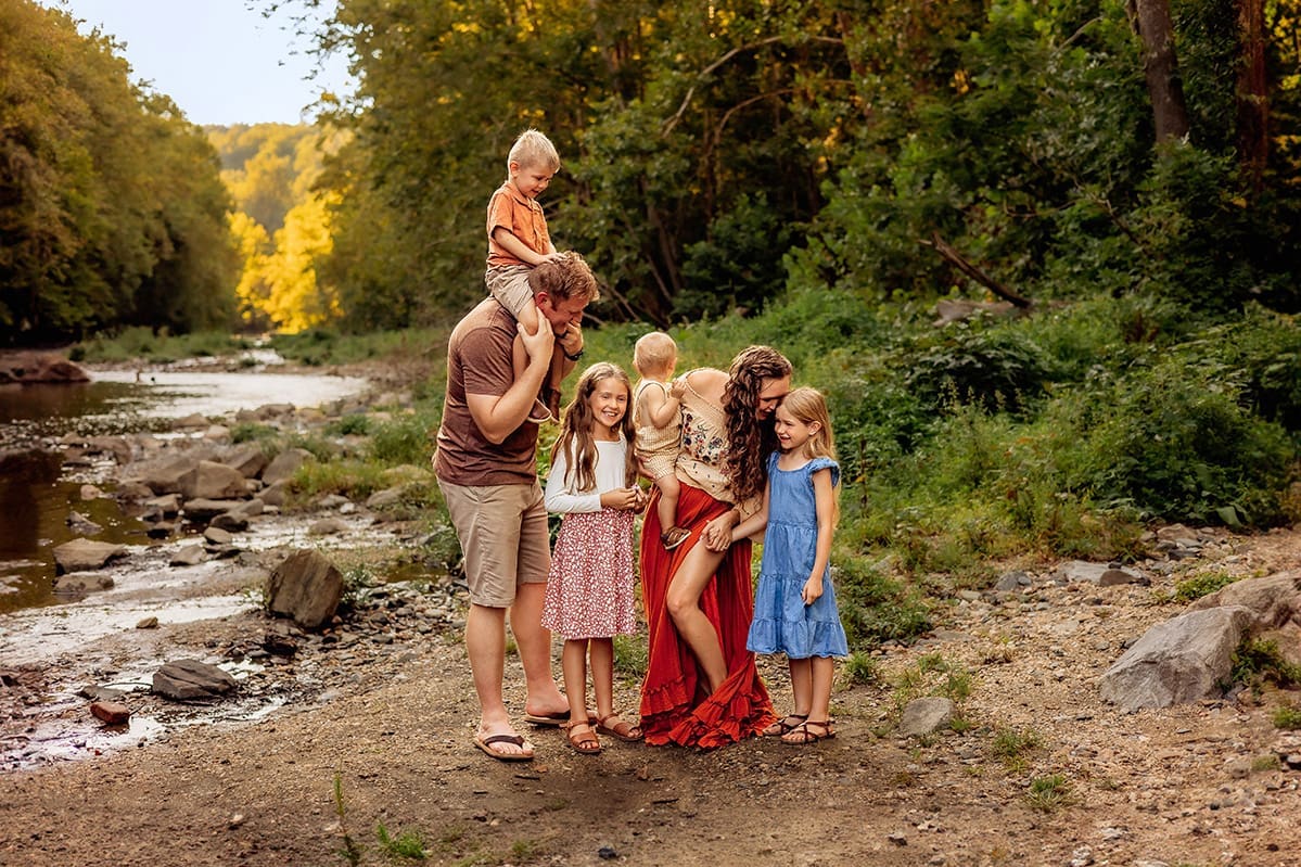 candid family photo session by a scenic creek at golden hour. A mother in a flowy red skirt leans down to connect with her daughters, while the father carries their son on his shoulders, all laughing together. Natural, warm, and playful family photography capturing real moments in nature.