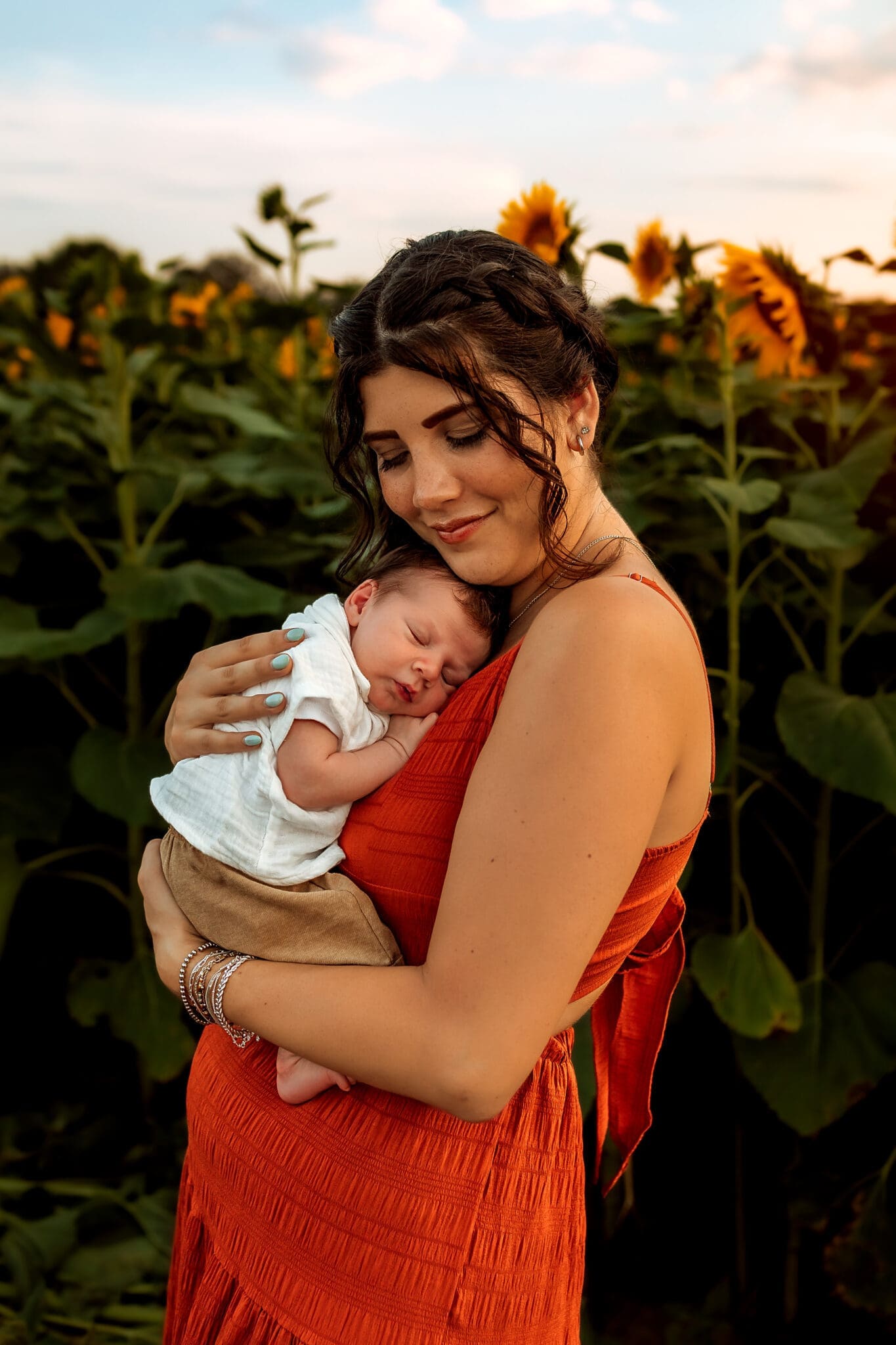 Mom holds newborn to chest and rests her cheek against him. They are standing in a sunflower field in Lisbon, MD.