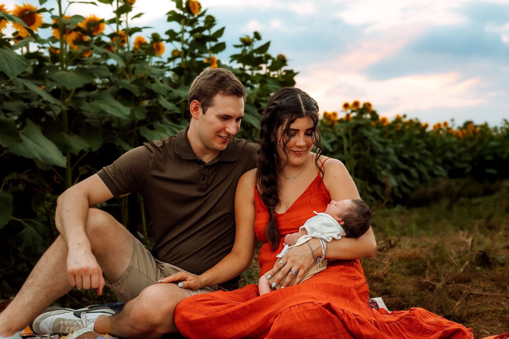 Mom and dad sit together in a sunflower field. Mom is holding their newborn. They both gaze down affectionately toward their baby.