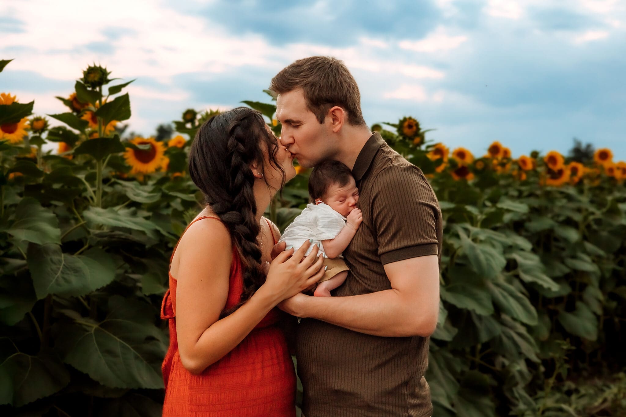 Mom and dad in a sunflower field. Dad holds newborn to his chest and leans over to give mom a kiss.