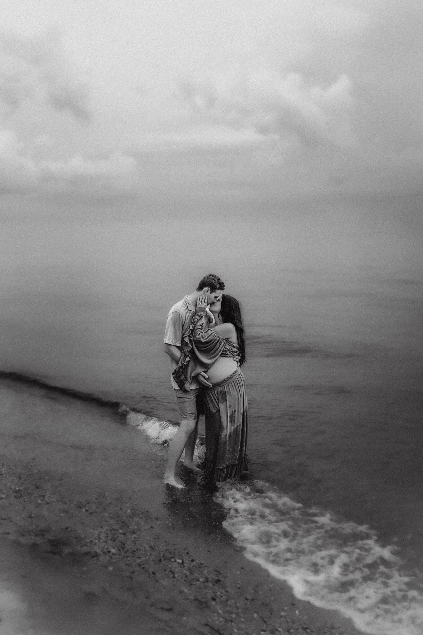 An expectant mother reaches up as her partner embraces her to pull him in for a kiss as the waves crash in on the edge of the Chesapeake Bay.