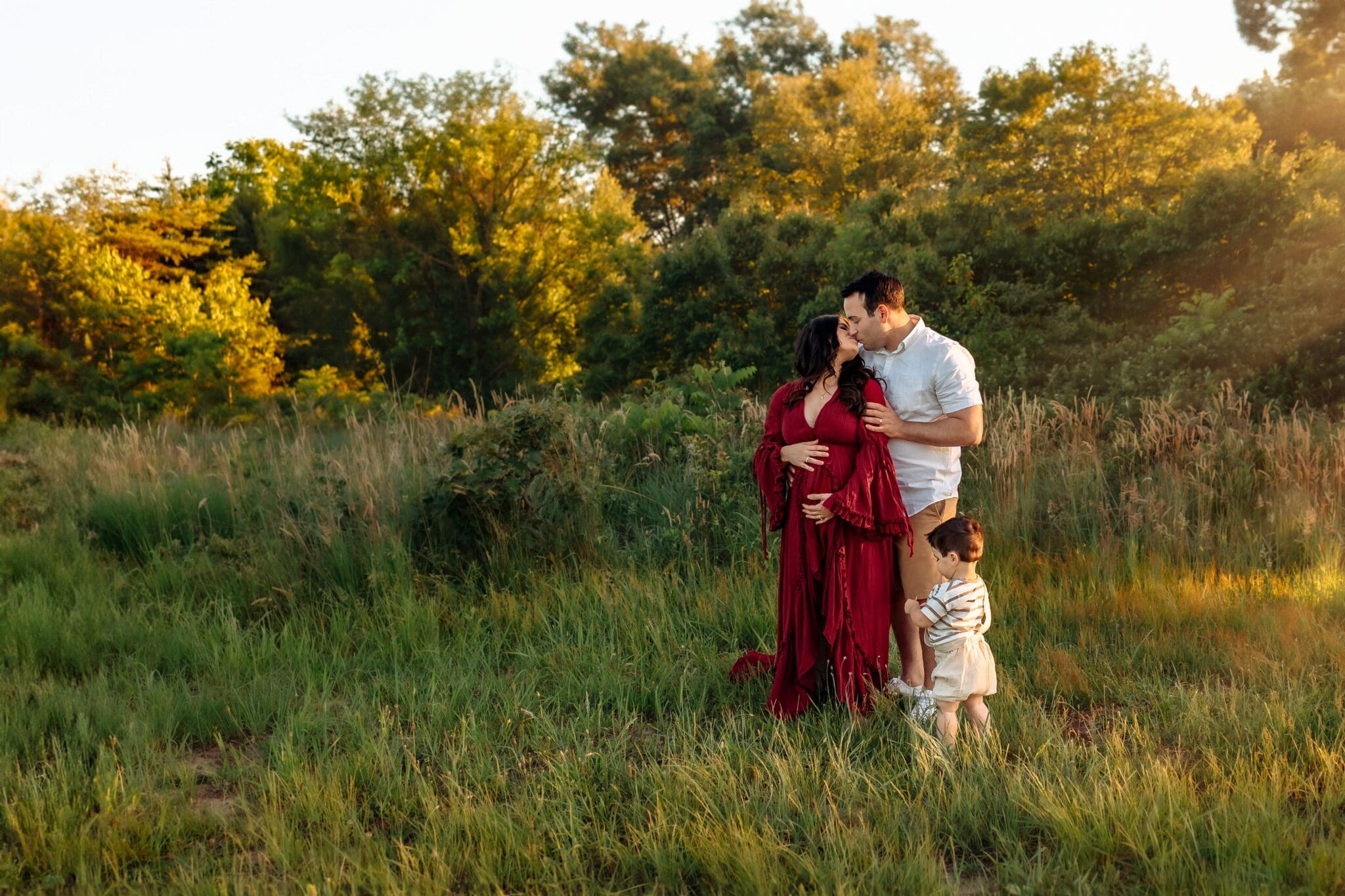 Outdoor maternity photographer Maryland, family of three embrace in a field of tall grasses, mother in a red dress