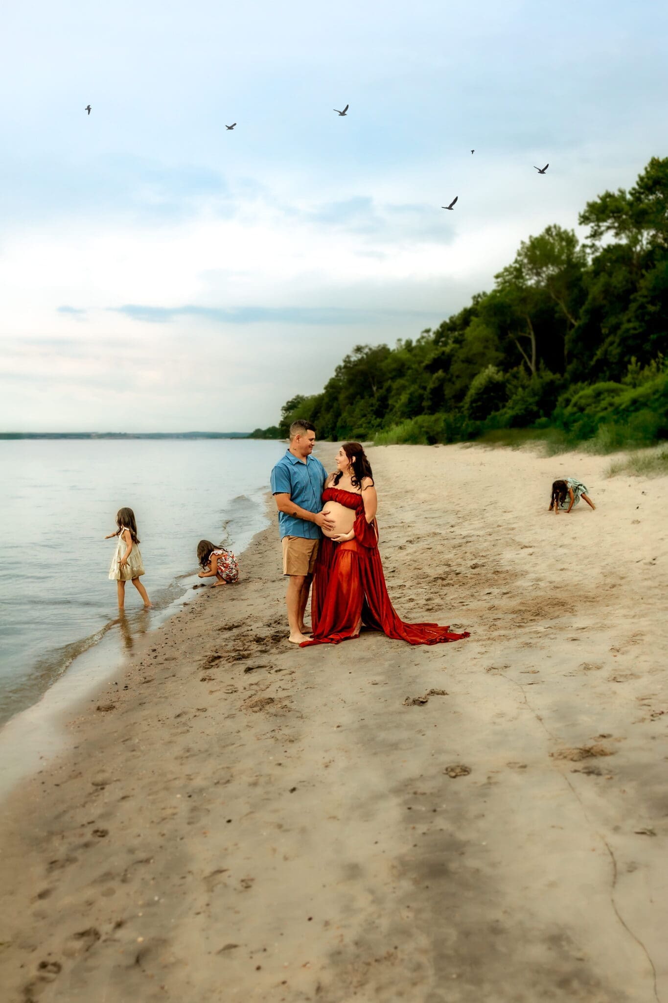 A family plays along the beach. Expectant mom in a rust dress standing with her husband on a wide open beach at dusk