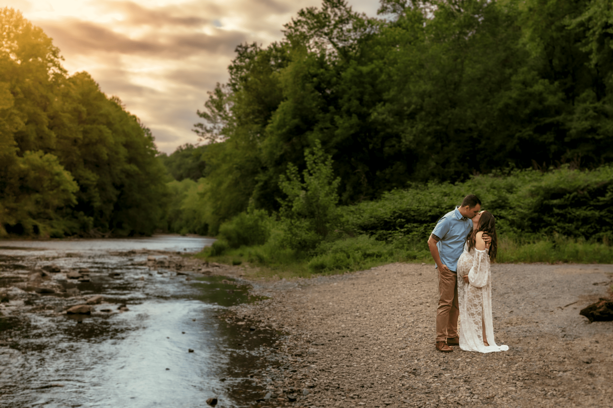 Pregnant woman in a white dress standing near a creek and kissing her partner in a wooded setting